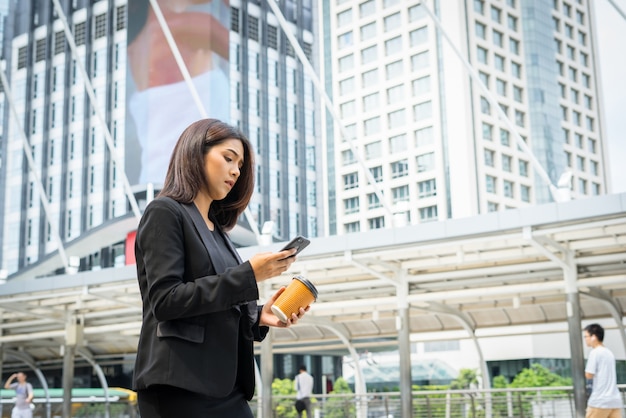 Mujer de negocios usando teléfono con café en la mano caminando en la calle con edificios de oficinas en el fondo