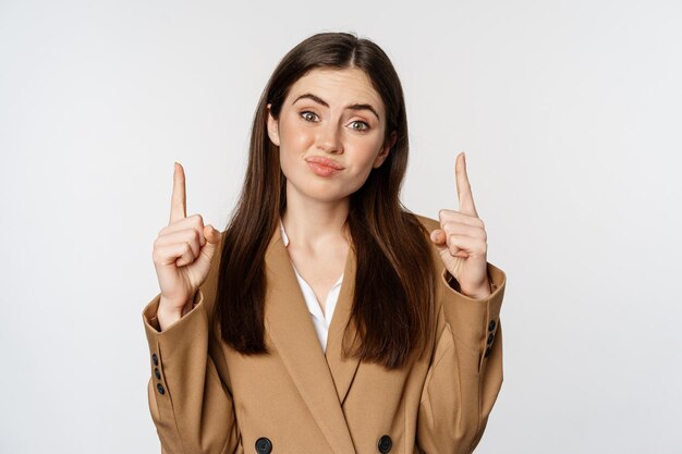 Mujer de negocios vacilante haciendo muecas de mal humor y señalando con el dedo hacia arriba con la cara decepcionada de pie en...