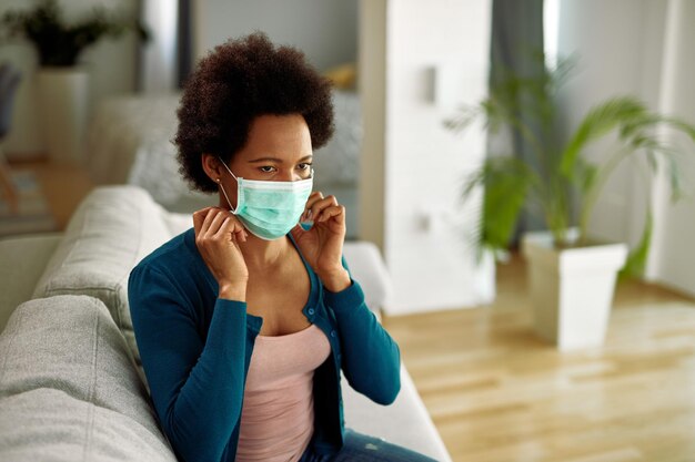 Mujer negra ajustando una máscara protectora en la cara mientras se relaja en la sala de estar