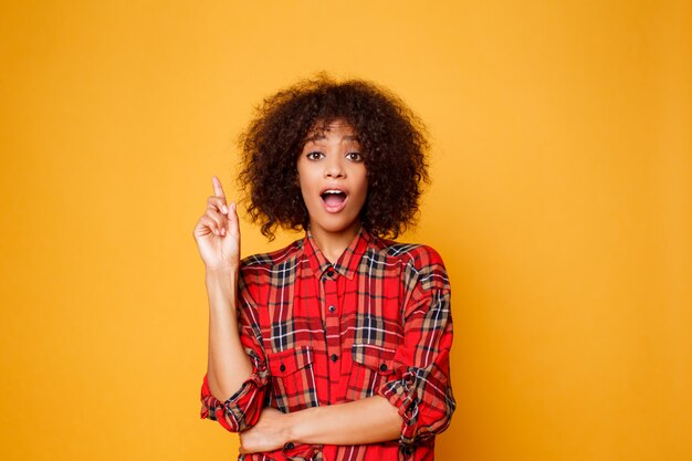 Mujer negra americana alegre en la camisa roja que mira en cámara y que señala el dedo para arriba en el espacio de la copia aislado sobre fondo anaranjado.