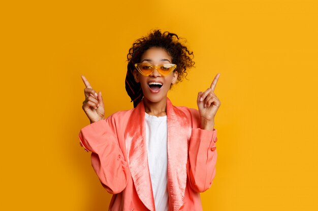 Mujer negra con cara graciosa apuntando hacia arriba. Con una chaqueta rosa casual. Fondo amarillo