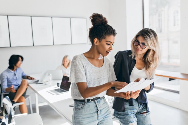 Mujer negra delgada en jeans explicando algo a la colega femenina europea mientras el hombre asiático habla con una joven rubia. Retrato de gerentes de empresa internacional resolviendo problemas laborales.