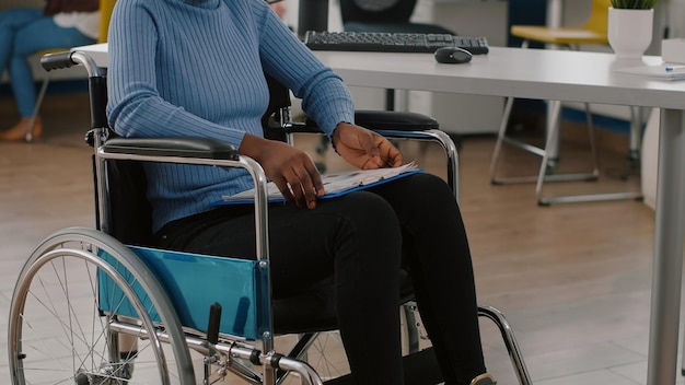 Mujer negra con discapacidad, inválido, discapacitado paralizado discapacitado sentado inmovilizado en silla de ruedas con portapapeles con documentos financieros. Gerente africano que trabaja en la sala de la oficina de negocios