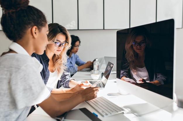 Mujer negra especialista en informática sentada al lado de una computadora moderna con pantalla negra y hablando con amigos. Retrato interior de jóvenes ocupados que trabajan en una empresa internacional.