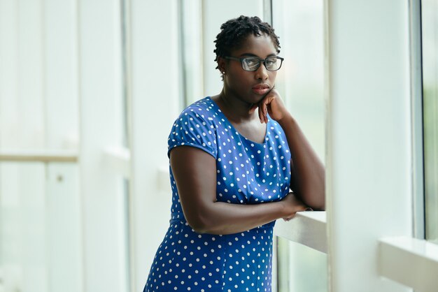 Mujer negra con gafas y vestido de lunares de pie junto a la ventana con la cabeza apoyada en la mano