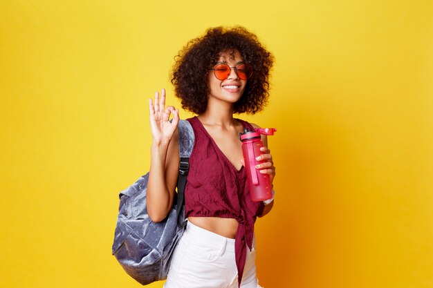 Mujer negra juguetona feliz en elegante traje de verano con signo de la paz posando en estudio sobre fondo amarillo. Sosteniendo una botella de agua. Peinado Afro. Estilo de vida saludable.