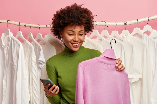 La mujer negra positiva elige un suéter para comprar, sostiene una percha con un cuello alto de color púrpura, un teléfono móvil en la otra mano