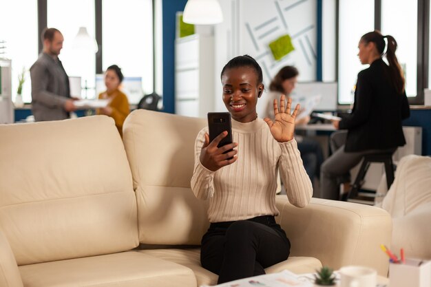 Mujer negra saludando a la cámara explicando los informes financieros al administrador remoto en la videollamada con teléfono inteligente con auriculares sentado en el sofá