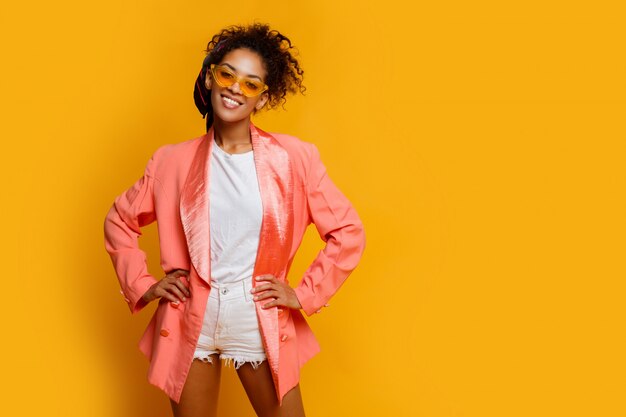 Mujer negra sonriente confiada en la presentación elegante de la chaqueta rosada interior en fondo amarillo.