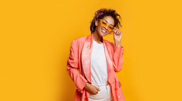 Mujer negra sonriente confiada en la presentación elegante de la chaqueta rosada interior en fondo amarillo.