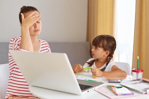 Mujer nerviosa cansada con camisa casual a rayas que está cansada para explicar la tarea del hogar, está agotada, cubriendo los ojos con la palma, niño mirando a la madre, educación en línea.