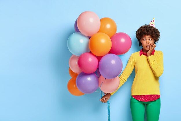 Mujer nerviosa perpleja sostiene globos multicolores mientras posa en un suéter amarillo