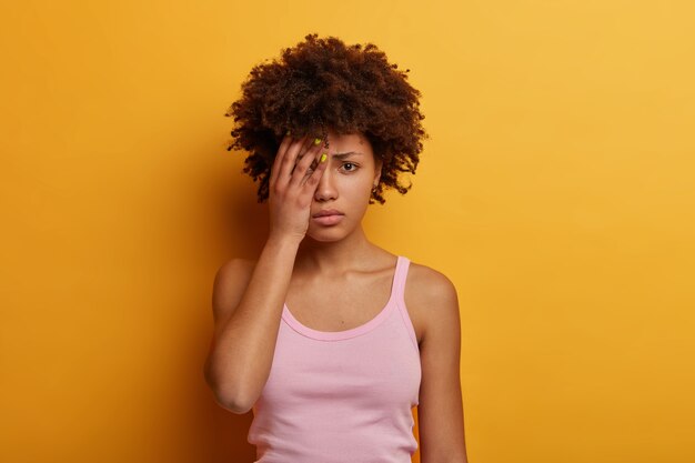 Mujer ofendida y abatida que va a llorar, se cubre la mitad de la cara, se ve triste, se siente decepcionada, siendo lastimada por alguien, vestida de manera informal, posa contra la pared amarilla. Emociones tristes