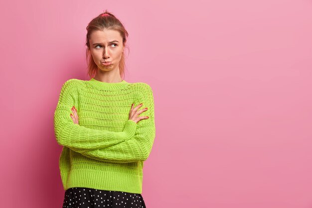 La mujer ofendida de mal humor se para con las manos cruzadas, frunce los labios, se siente ofendida, mira por debajo de la frente, usa un suéter verde, se para sobre una pared rosa