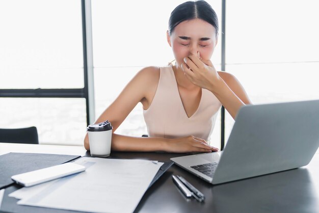 Mujer de oficina asiática enferma mala salud con alergia climática mientras trabaja con una computadora portátil en la oficina