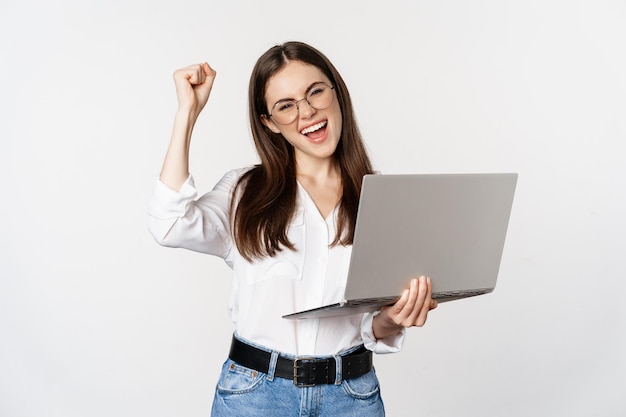 Mujer de oficina entusiasta, mujer de negocios sosteniendo una computadora portátil y gritando de alegría, celebrando y regocijándose, de pie sobre fondo blanco