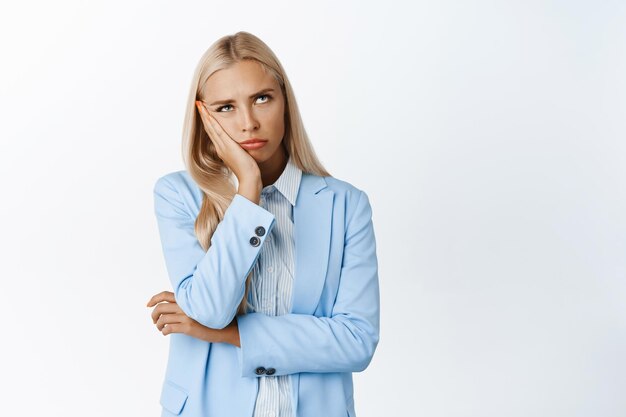 Mujer de oficina joven aburrida o molesta en traje pone los ojos en blanco y la cara inclinada en la mano de pie molesta contra el fondo blanco