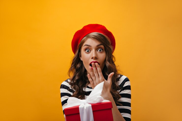 Foto gratuito la mujer de ojos azules parece sorprendida y se hace presente. chica sorprendida con pelo rizado en boina roja y camisa a rayas tiene caja de regalo.