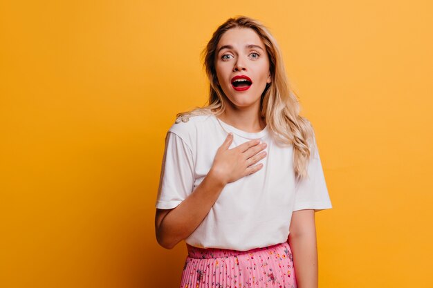 Mujer de ojos azules sorprendida de pie sobre la pared amarilla. Espectacular chica rubia posando emocionalmente