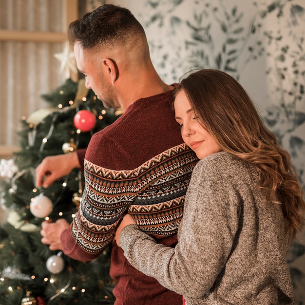 Mujer con los ojos cerrados que abrazan al hombre de atrás en suéteres cerca del árbol de Navidad