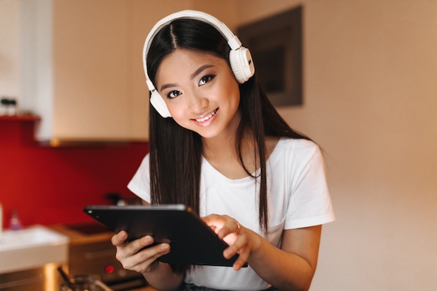 Mujer de ojos marrones con auriculares masivos mira al frente, sonríe y sostiene la tableta