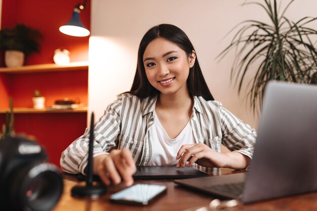 Mujer de ojos marrones en camisa a rayas sonriendo y posando en el lugar de trabajo con smartphone y portátil