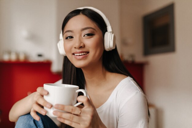 Mujer de ojos marrones en top blanco sosteniendo la taza y posando con una sonrisa