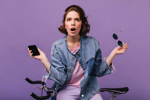 Mujer de ojos oscuros preocupada posando con teléfono. Foto interior de dama blanca emocional en chaqueta de mezclilla de pie junto a la bicicleta.