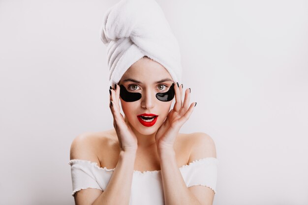 Mujer de ojos verdes con lápiz labial rojo cuida la piel debajo de los ojos. Retrato de modelo después de la ducha en la pared blanca.