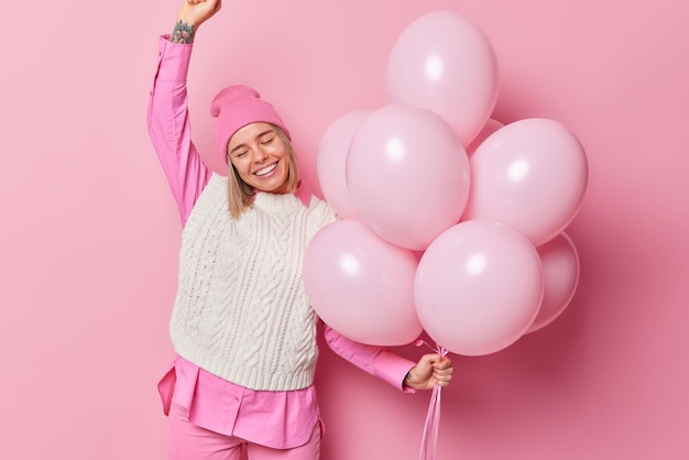 Una mujer optimista y complacida viste ropa de moda, baila al ritmo de la música en la fiesta, tiene poses de humor festivo contra un fondo rosa con globos inflados, celebra cumpleaños con amigos, concepto de vacaciones