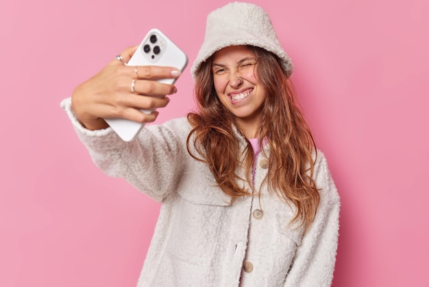 La mujer optimista y despreocupada positiva guiña los ojos, muerde los labios y posa en la cámara del teléfono inteligente, toma selfie, usa chaqueta blanca de moda, sombrero de piel, hace clic para hacer una foto de sí misma aislada sobre fondo rosa