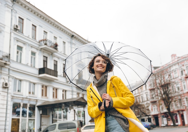 Mujer optimista en impermeable amarillo y gafas divirtiéndose mientras camina por la ciudad bajo un gran paraguas transparente durante el frío día lluvioso