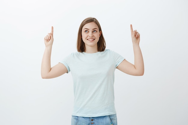 Mujer optimista intrigada y alegre con amplia sonrisa curiosa apuntando y mirando hacia arriba