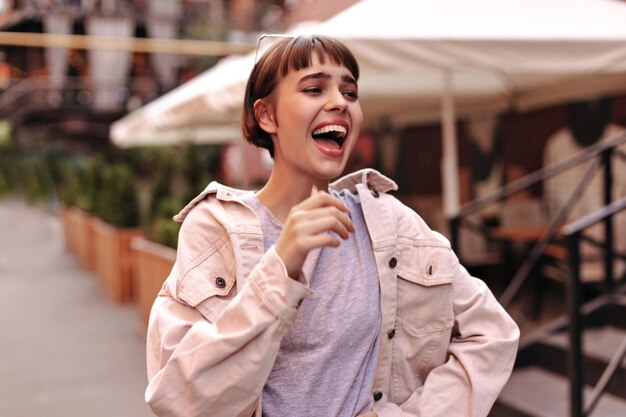 Mujer optimista con peinado corto en traje ligero riéndose de la calle Mujer morena alegre en chaqueta rosa de mezclilla posando afuera