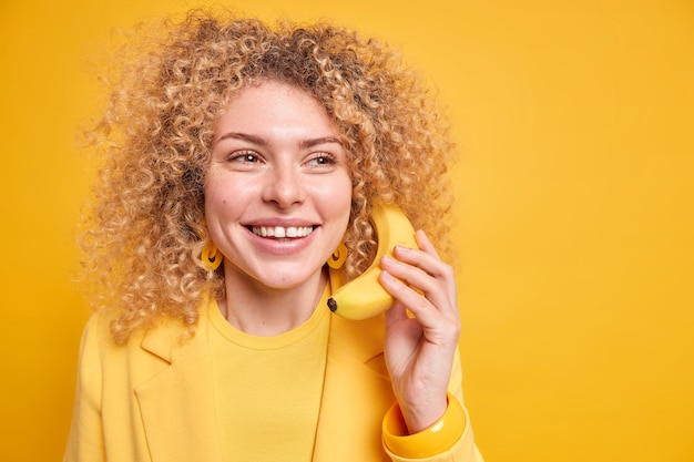 La mujer optimista de pelo rizado sonríe agradablemente sostiene el plátano cerca de la oreja como si una conversación telefónica mirara hacia otro lado con expresión soñadora aislada sobre el espacio de copia de la pared amarilla.