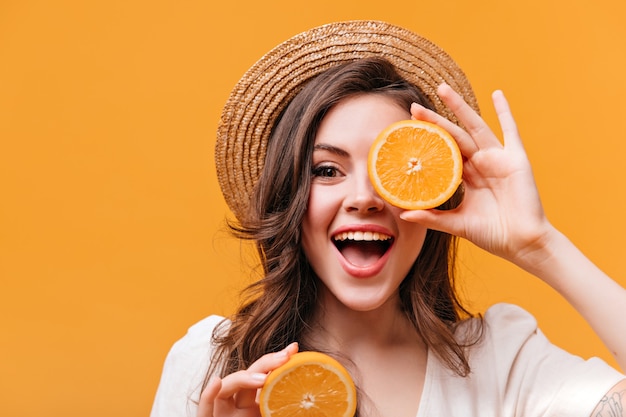 Mujer optimista con sombrero de paja se cubre los ojos con naranja y sonríe mientras mira a la cámara.