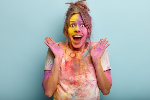 La mujer optimista sorprendida pasa un festival impresionante de colores, sonríe y extiende las manos hacia los lados, sucia con polvos de colores, reacciona feliz