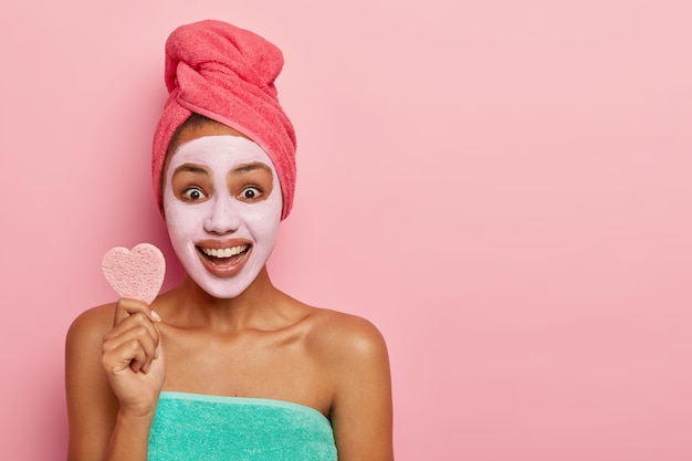 La mujer optimista sostiene una pequeña esponja suave para tratamientos faciales, se para envuelta en una toalla, sonríe ampliamente, se aplica una mascarilla de arcilla fresca para limpiar la cara y la piel sana. Copiar espacio para texto