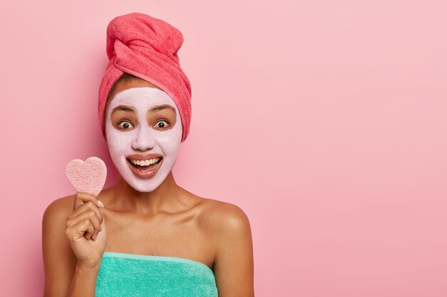 La mujer optimista sostiene una pequeña esponja suave para tratamientos faciales, se para envuelta en una toalla, sonríe ampliamente, se aplica una mascarilla de arcilla fresca para limpiar la cara y la piel sana. Copiar espacio para texto