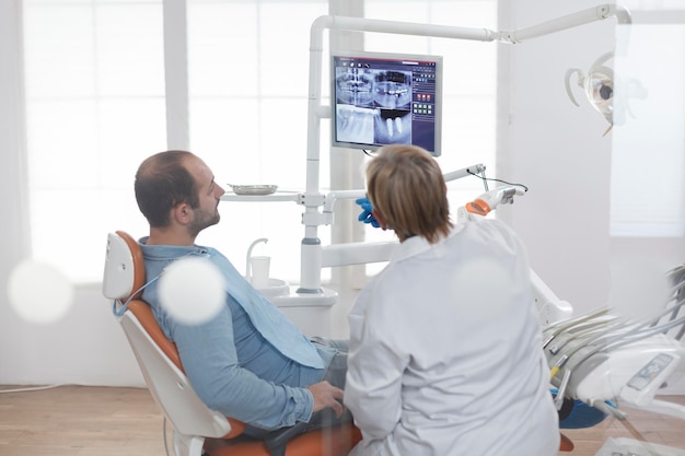 Mujer ortodoncista de alto nivel con mascarilla facial explicando la radiografía de odontología al paciente discutiendo el tratamiento oral para prevenir infecciones durante el examen médico en el consultorio dental. Concepto de estomatología