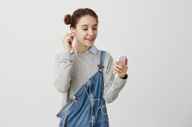 Mujer en overol de mezclilla estar relajado con smartphone viendo videos de internet. Chica adulta escuchando música a través de auriculares con sonrisa. Concepto de tecnología