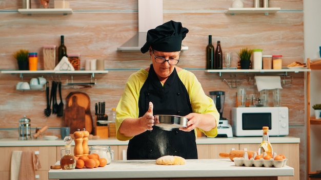 Foto gratuita mujer panadero con harina tamiz metálico preparando tartas caseras. feliz chef anciano con bonete preparando ingredientes crudos para hornear pan tradicional espolvoreado, tamizado en la cocina.