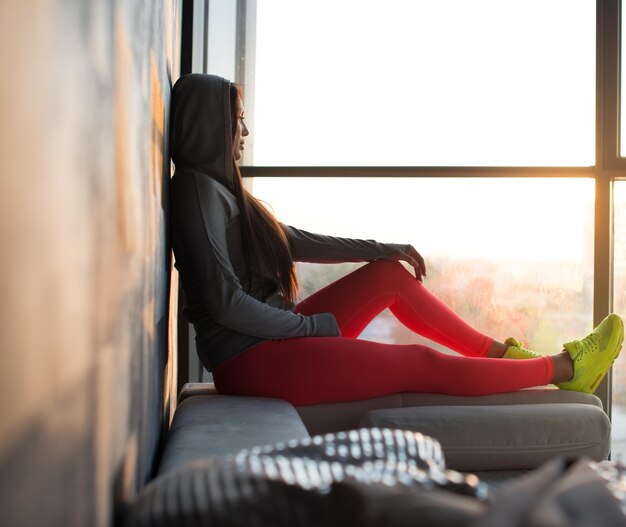 Una mujer con pantalones deportivos rojos y choes amarillos sentada cerca de la ventana y mirando hacia afuera.