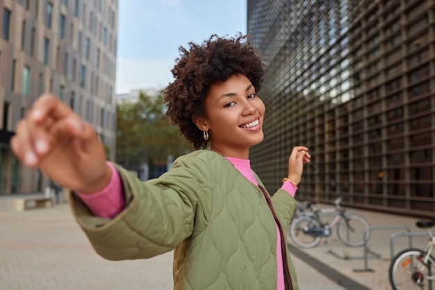 Foto gratuita mujer pasea por la ciudad y baila mantiene los brazos a los lados disfruta de un buen día camina en el centro de la ciudad entre los edificios modernos de la ciudad usa chaqueta de buen humor