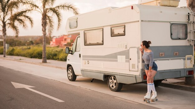 Mujer patinadora de pie detrás de la caravana en la carretera mirando a escondidas