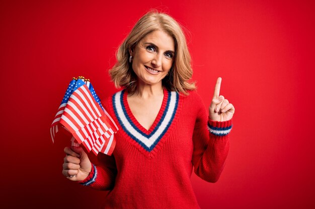 Mujer patriótica rubia de mediana edad sosteniendo banderas de estados unidos celebrando el día de la independencia sorprendida con una idea o pregunta señalando con el dedo con la cara feliz número uno