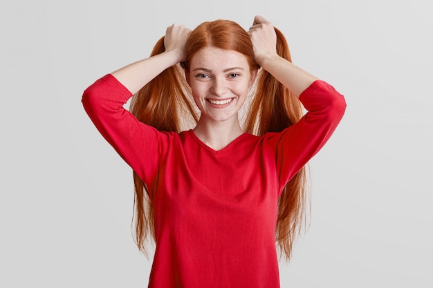 La mujer pecosa pelirroja positiva tiene el pelo largo, hace cola de caballo, se complace en posar ante la cámara, usa un suéter rojo informal