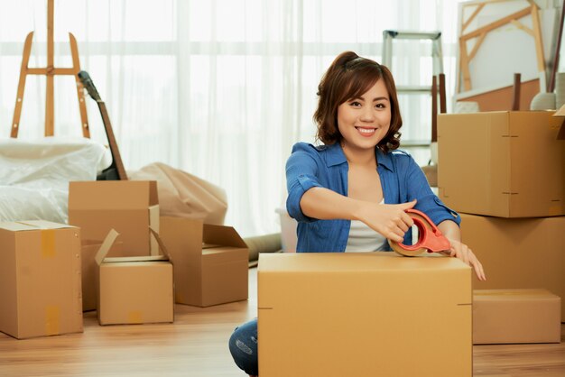 Mujer pegando adhesivo a la caja del paquete sonriendo a la cámara sentada en el suelo