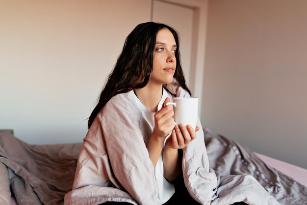 Mujer con peinado largo y ondulado está sentada en la cama con café después de despertarse por la mañana y mirando a la ventana Retrato de una joven soñadora linda descansando en el dormitorio por la mañana