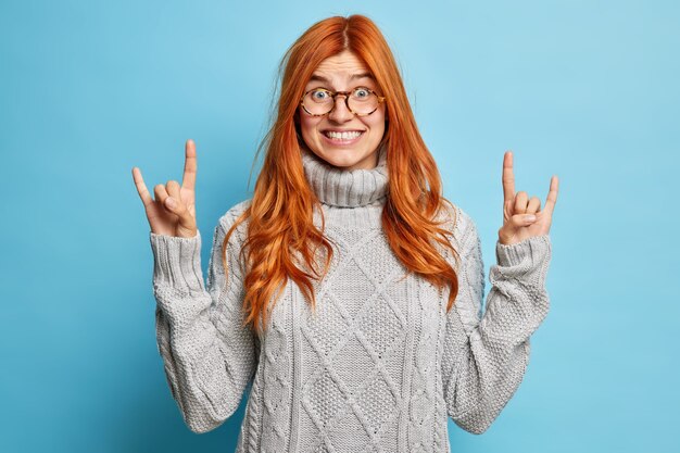 Mujer pelirroja alegre complacida en espectáculos muestra rock n roll signo de mano gesto de cuerno de diablo sonríe ampliamente muestra dientes blancos usa suéter de invierno.